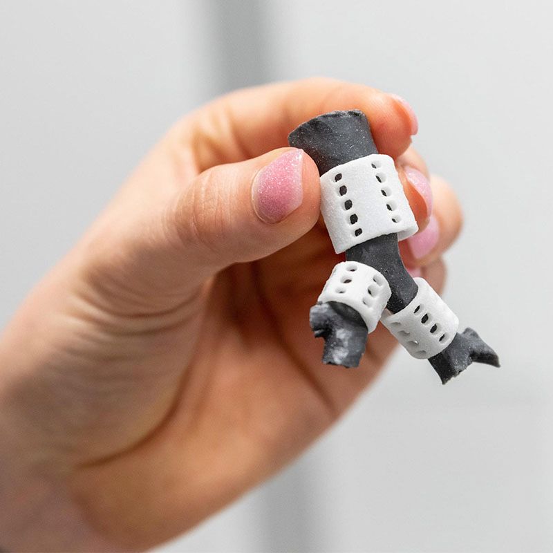 A hand holding a small 3D‑printed medical prototype with white perforated brackets attached to a dark cylindrical structure.
