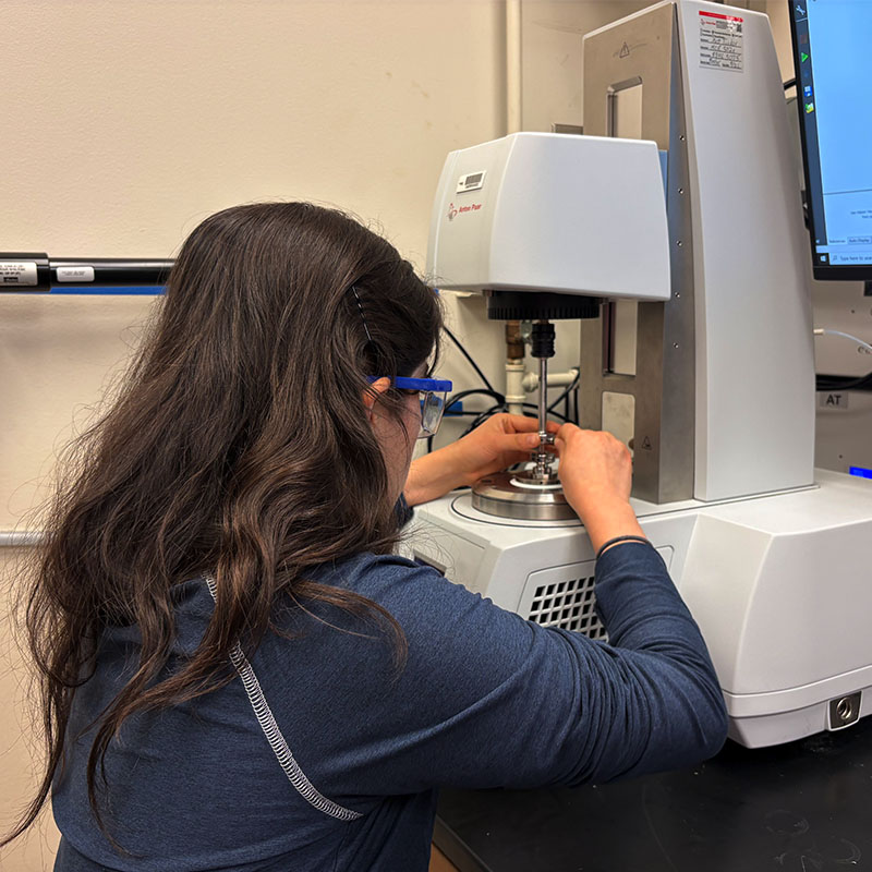 A researcher adjusting a sample on a mechanical testing instrument at a lab bench while wearing safety glasses.