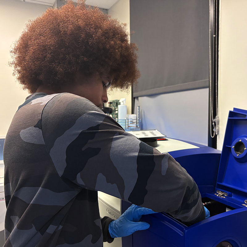 A researcher wearing gloves retrieves materials from a blue laboratory instrument used for biopolymer processing.