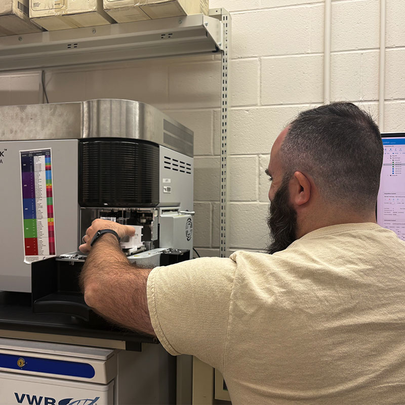 A researcher loading samples into a flow cytometry instrument at a laboratory bench, with a monitoring screen visible nearby.