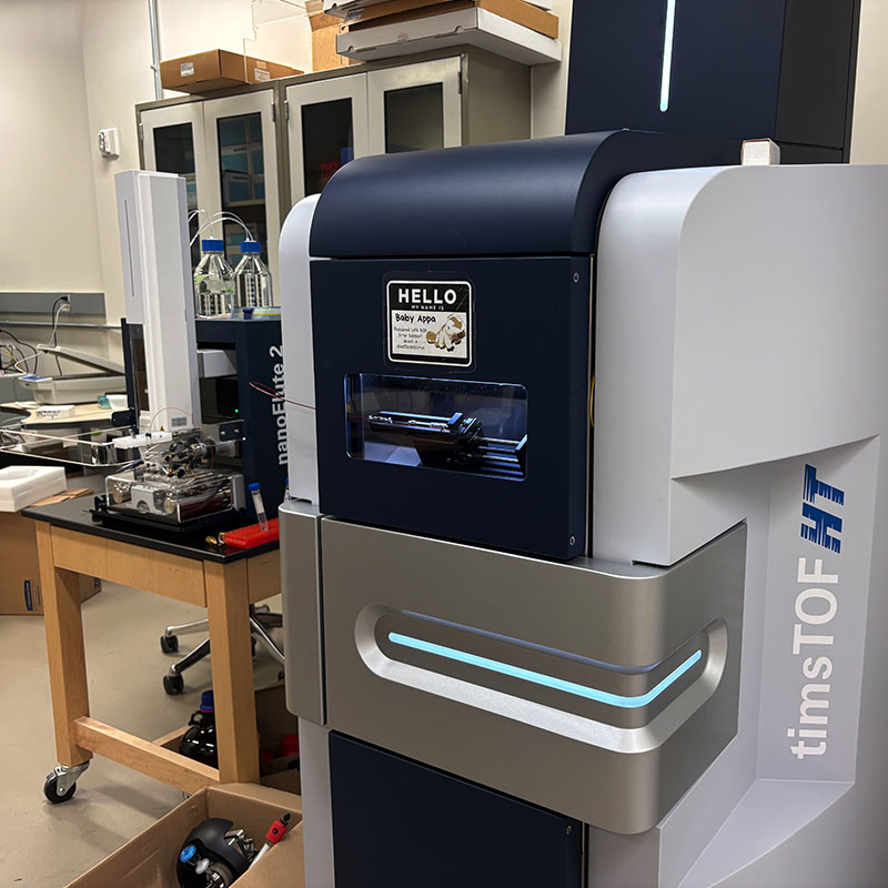 A timsTOF mass spectrometry instrument in a proteomics laboratory, with tubing, sample components, and nearby lab equipment visible.