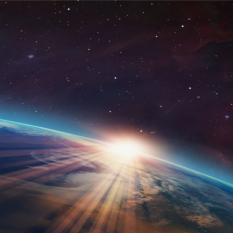 Sunlight rises over Earth's curved horizon as seen from space, casting bright rays across the planet's cloud-covered surface against a star-filled sky