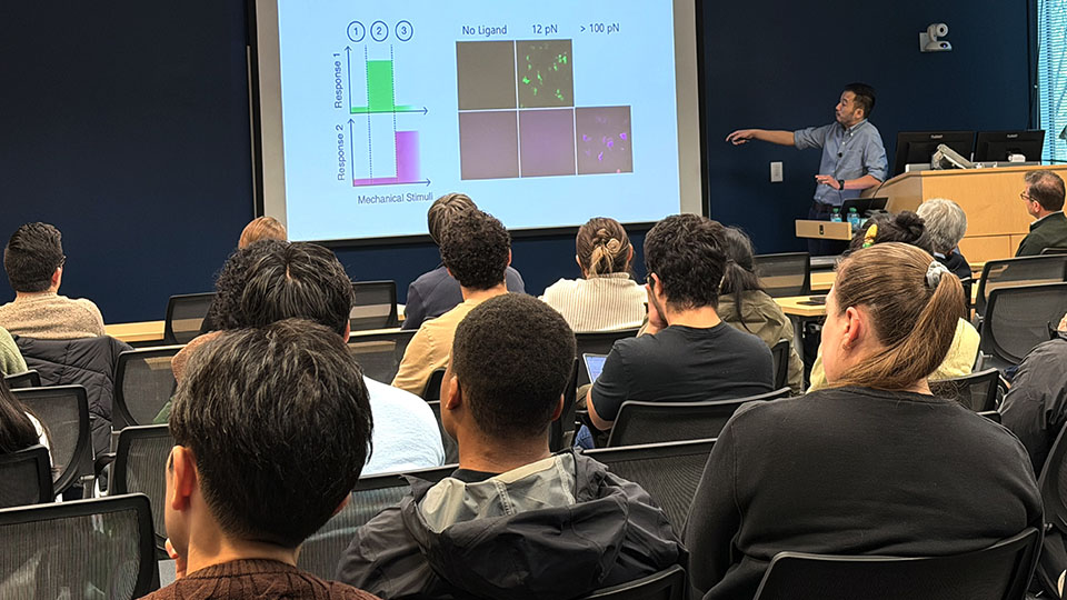 Audience listening to a presenter giving a scientific talk with data charts and fluorescent cell images displayed on a projection screen.