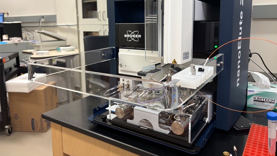 A benchtop mass spectrometry system with tubing, vials, and a clear protective enclosure in a laboratory.