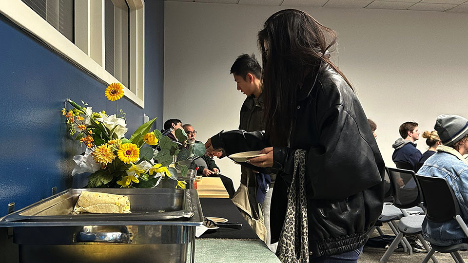 A woman wearing a black leather jacket puts food from a buffet on her plate while a man in the background does the same.