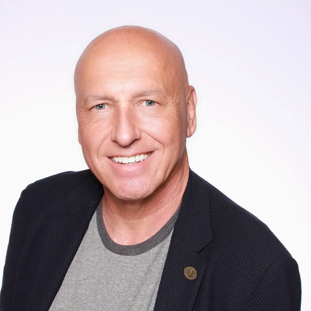 Professional headshot of a smiling bald man with light skin, wearing a dark textured blazer over a gray crew-neck shirt. He is facing the camera against a light gray background, with a friendly and approachable expression. The portrait is cropped from the chest up and evenly lit, creating a clean, professional appearance.